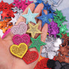 A hand holds a set of five colorful glitter heart and star iron-on embroidered patches in various shades of pink, blue, green