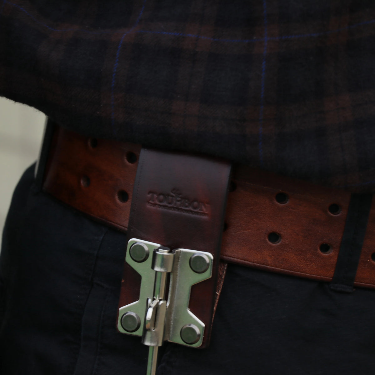 A close-up of a brown leather belt with a bronze buckle and a steel J-hook attached to a black garment.