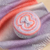 A roll of colorful gradient cake yarn in pink and purple shades displayed on a purple knitted fabric.