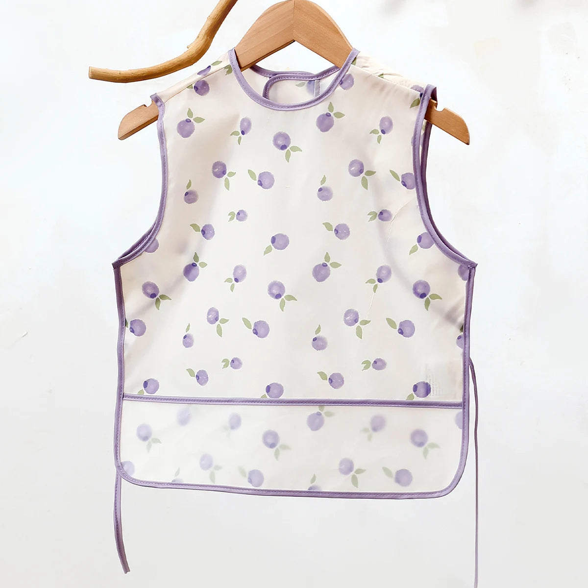 A sleeveless white baby feeding bib with a purple fruit pattern hanging on a wooden hanger.