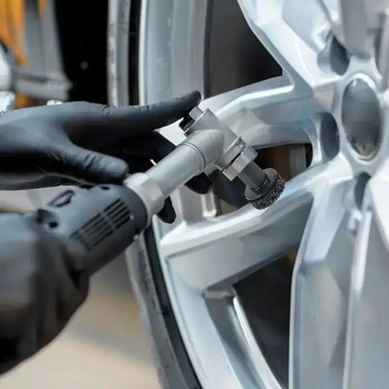 A gloved hand uses a drill attachment to clean or polish the hub of a silver car wheel.