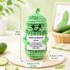 Handmade crochet green pickle-shaped pocket hug doll with a smiling face resting on a woven mat.