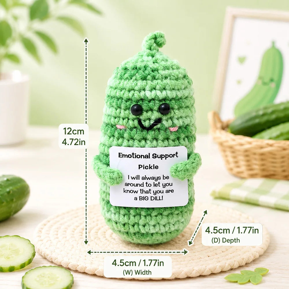 Handmade crochet green pickle-shaped pocket hug doll with a smiling face resting on a woven mat.