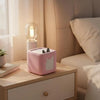 A pink silicone protective case for a Toniebox 2 audio player sitting on a wooden surface next to a lamp.