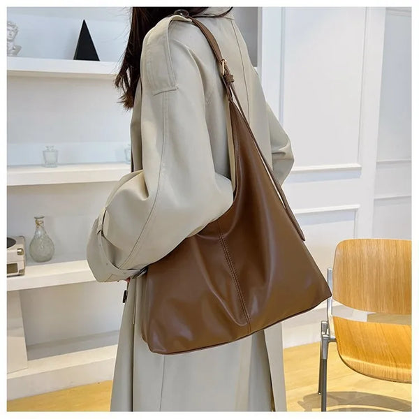 A woman is holding a tan leather retro tote bag in a light beige coat against a white background.