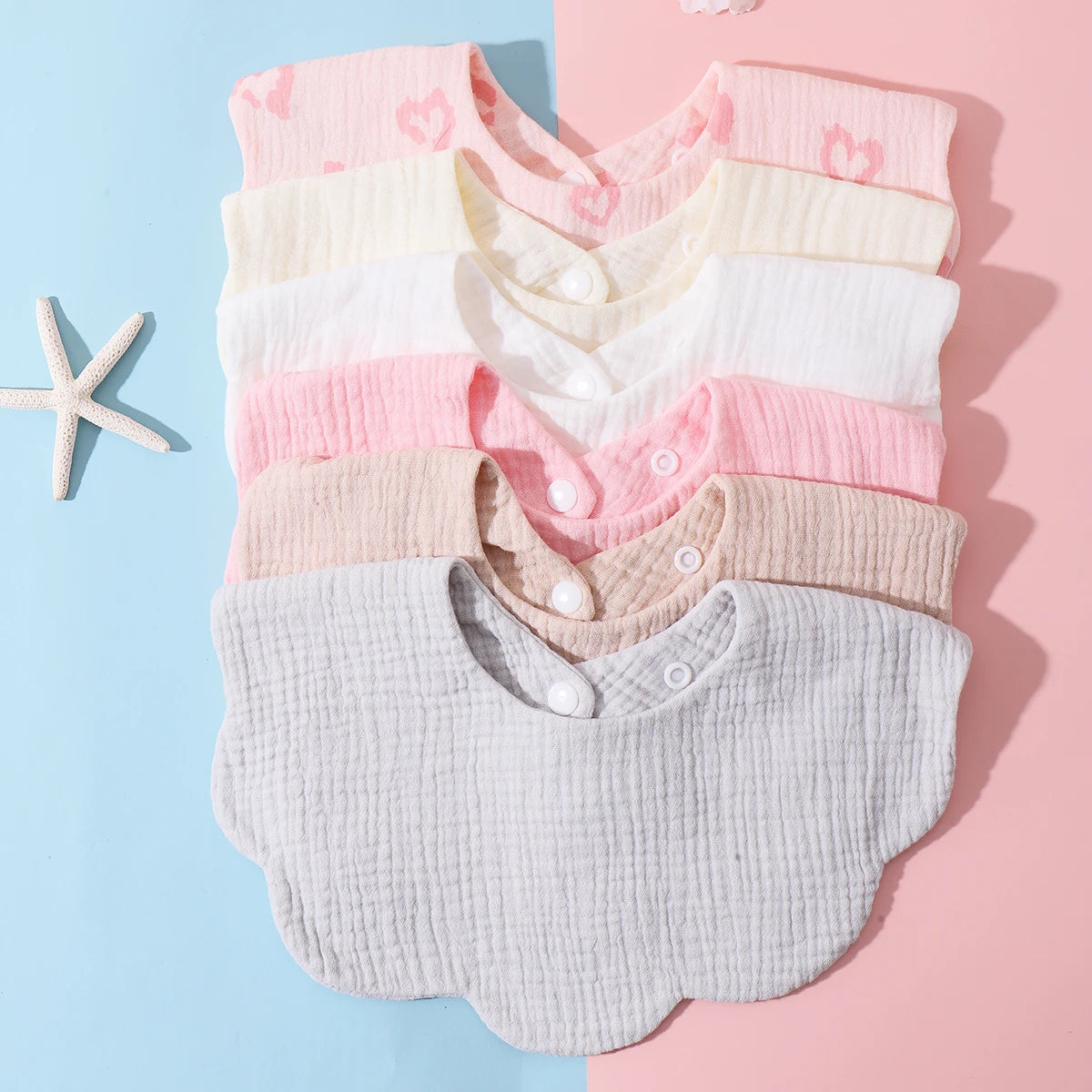 A stack of three soft cotton baby bibs in pastel pink and cream colors displayed on a light blue and pink background.