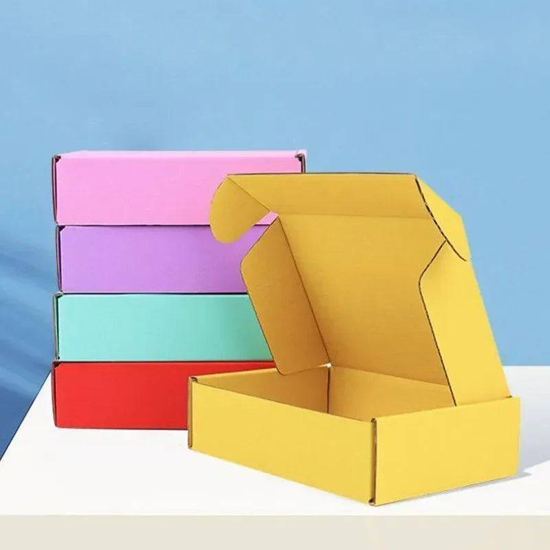 Ein Stapel bunter Wellpappkartons in rosa, lila und türkis neben einer offenen gelben Box.