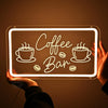 A hand holds a glowing neon sign shaped like coffee beans and a bar, illuminating the text Coffee Bar.
