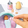 A hand holds a pink baby doll accessory within a purple bowl, next to a yellow teapot and a pink formula container.
