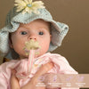 A baby wearing a light blue knitted hat with a flower, holding a silicone teether toy shaped like a flower and grape.