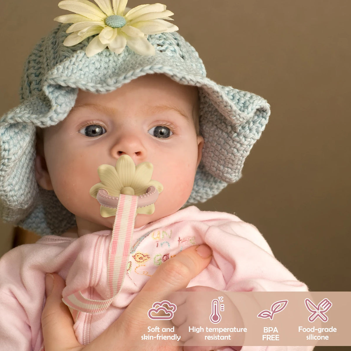 A baby wearing a light blue knitted hat with a flower, holding a silicone teether toy shaped like a flower and grape.