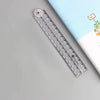 A transparent plastic ruler with a measuring tape and a circular marker is shown next to a notebook.