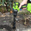 A hand holds a green garden auger with a black drill bit inserted into the soil.