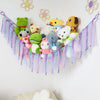 A bohemian handwoven corner toy storage net with colorful stuffed animals and tassels hanging on a white wall.