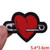 A red heart embroidered iron-on patch with a silver pin attached on a black fabric background.