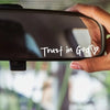 A hand holds a black vinyl sticker with the text Trust in God and a heart symbol, positioned against a blurred background.