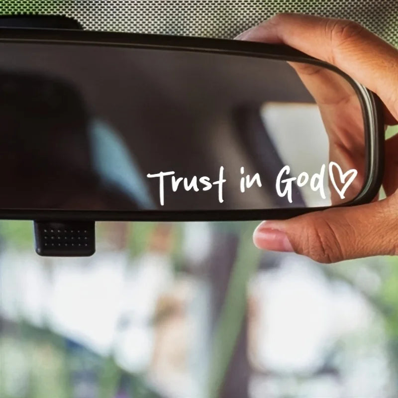 A hand holds a black vinyl sticker with the text Trust in God and a heart symbol, positioned against a blurred background.