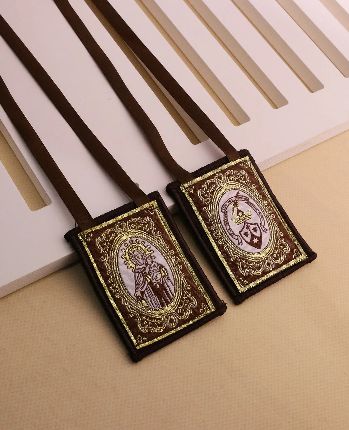 A brown leather scapular necklace with an ornate pendant featuring a religious figure on a dark fabric background.