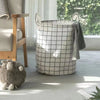 A large, white and black checkered laundry basket with handles sits on a light gray floor next to a wooden chair and a sheep.