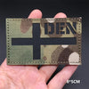 A rectangular tactical patch featuring a dark cross on a camouflage background with the letters DEN visible.
