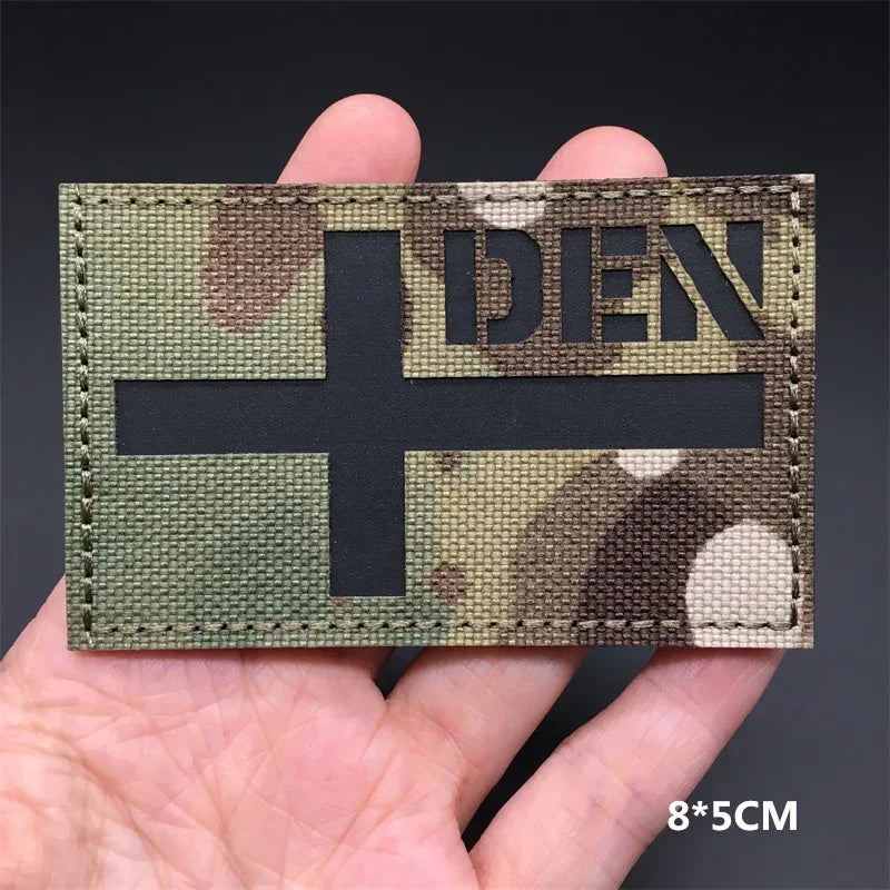 A rectangular tactical patch featuring a dark cross on a camouflage background with the letters DEN visible.