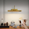 A gold cordless rechargeable picture light is shown above a wooden surface with decorative items.