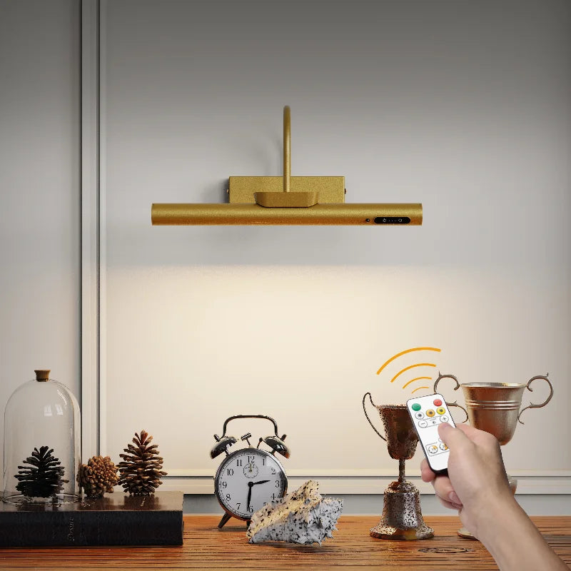 A gold cordless rechargeable picture light is shown above a wooden surface with decorative items.