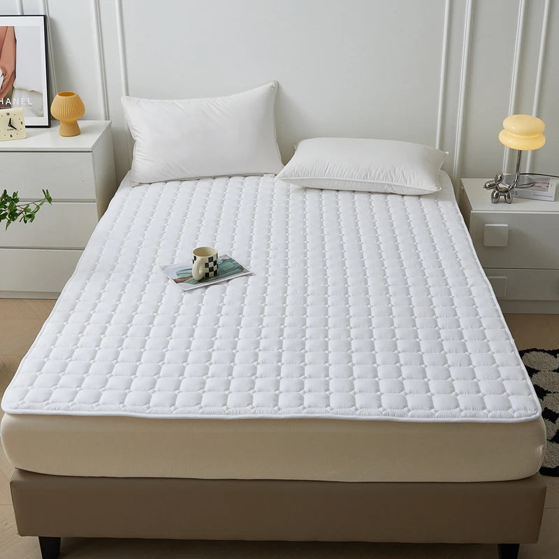 Weißes gestepptes Matratzenschoner liegt auf einem Bett neben Nachttischen und Kissen.