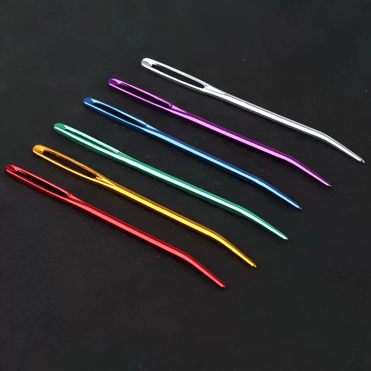 A collection of twenty large eye tapestry needles with curved tips in various colors laid on a dark surface.