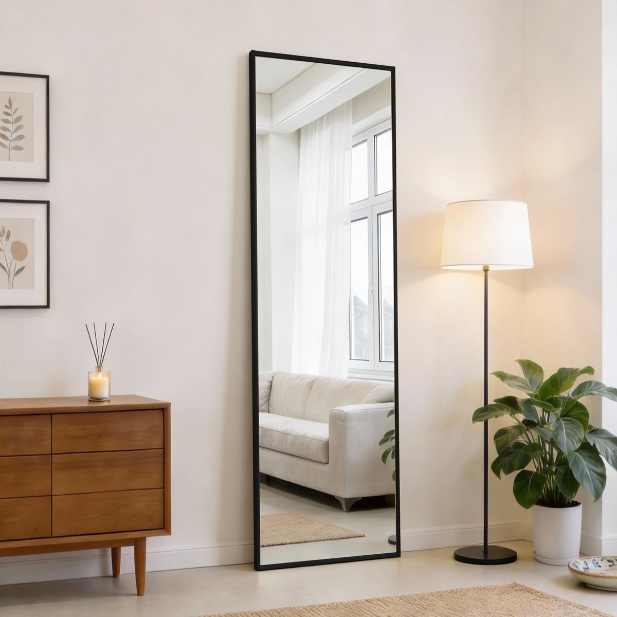A full length rectangular mirror with a black frame reflecting a bright window and interior decor.
