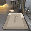 A rectangular beige bath mat with the word Bathroom embroidered on it, placed on a dark gray floor.