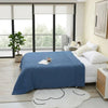 A deep blue pure cotton muslin throw blanket is displayed on a bed in a bright room with large windows.