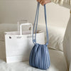 A light blue pleated faux leather crossbody bag is being held above a white paper shopping bag on a white sofa.