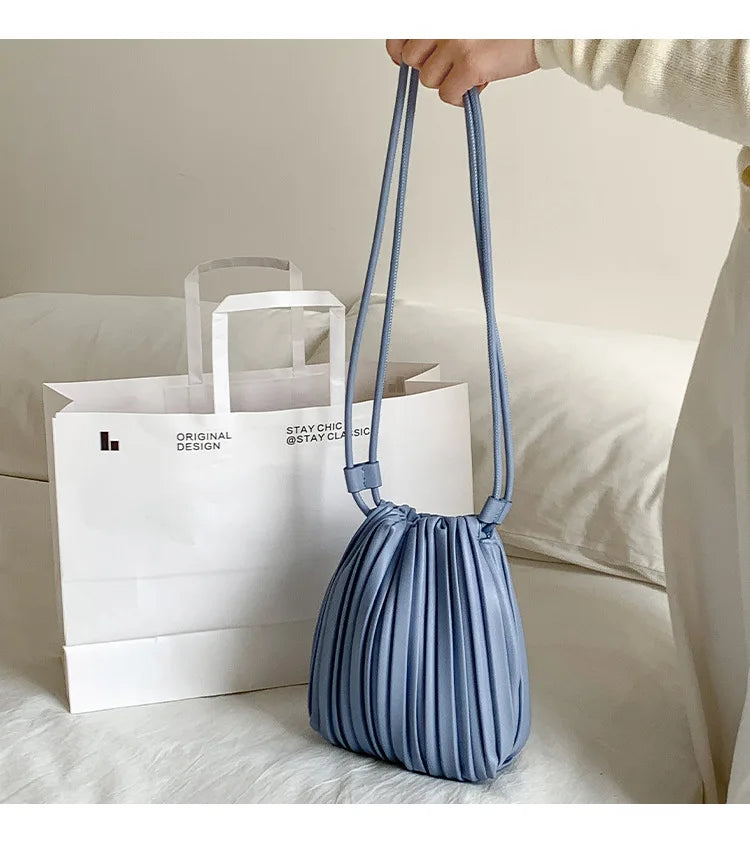 A light blue pleated faux leather crossbody bag is being held above a white paper shopping bag on a white sofa.