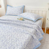 A light blue floral quilted bedspread with a pale blue trim is displayed on a bed in a bright bedroom.