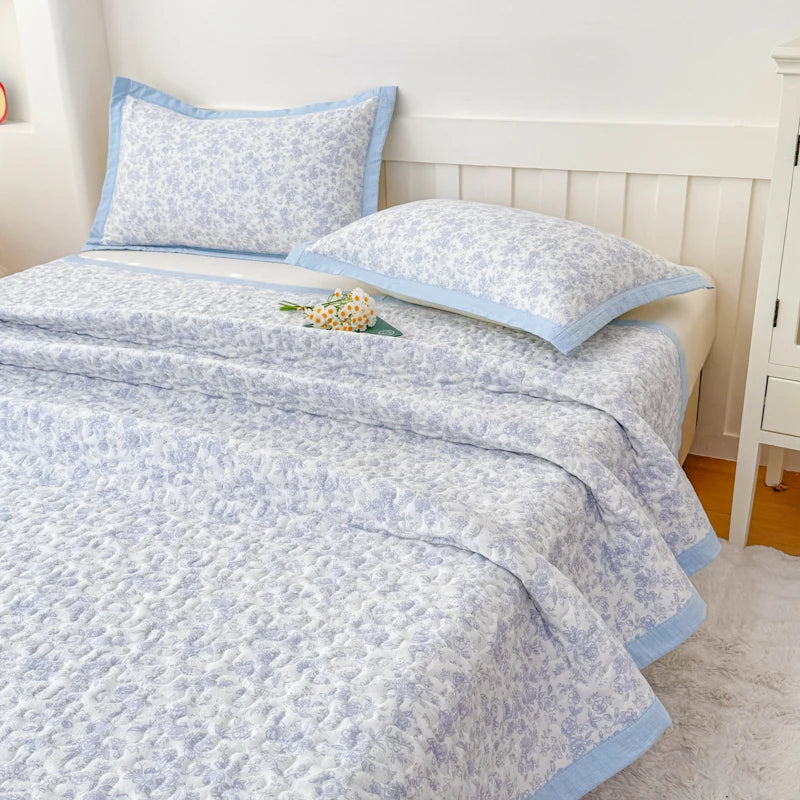 A light blue floral quilted bedspread with a pale blue trim is displayed on a bed in a bright bedroom.