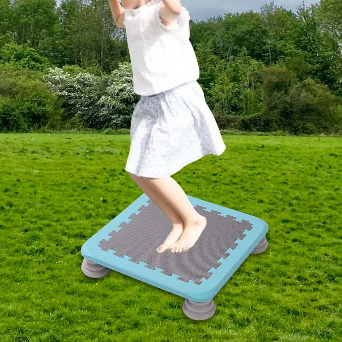 A child is standing on a blue mini trampoline mat outdoors in a grassy field.