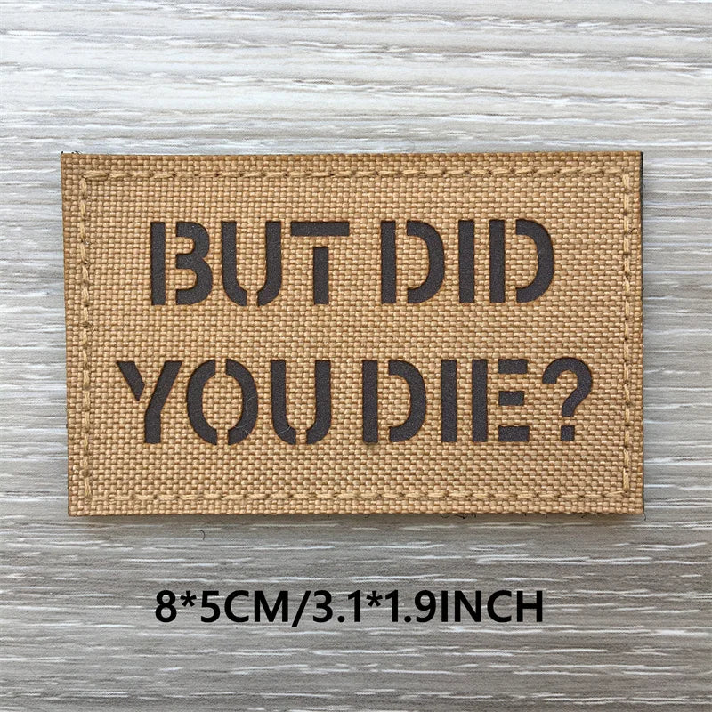 A rectangular tan woven patch with black text that reads BUT DID YOU DIE? on a gray wooden surface.