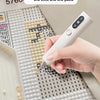 A hand holds a white electric diamond art pen with a vacuum suction feature, positioned over a grid of small white beads.