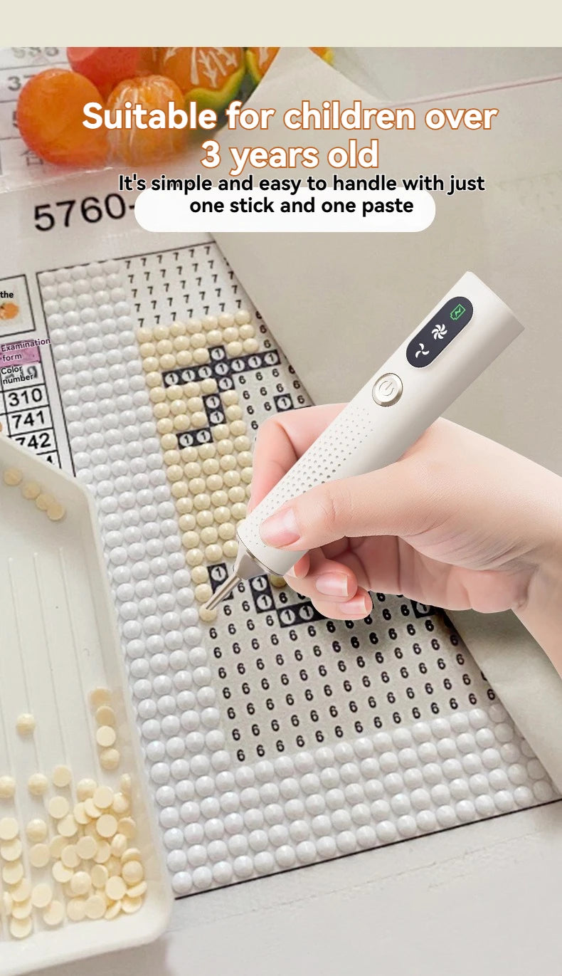 A hand holds a white electric diamond art pen with a vacuum suction feature, positioned over a grid of small white beads.