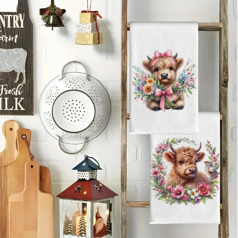 A set of two cute Highland cow kitchen towels featuring floral designs displayed on a wooden rack.