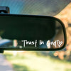 A close-up shot of a black rearview mirror showing the white text Trust in God with a heart symbol.