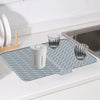 A silicone dish drying mat with a drainage ramp is placed on a sink next to drinking glasses and a storage container.