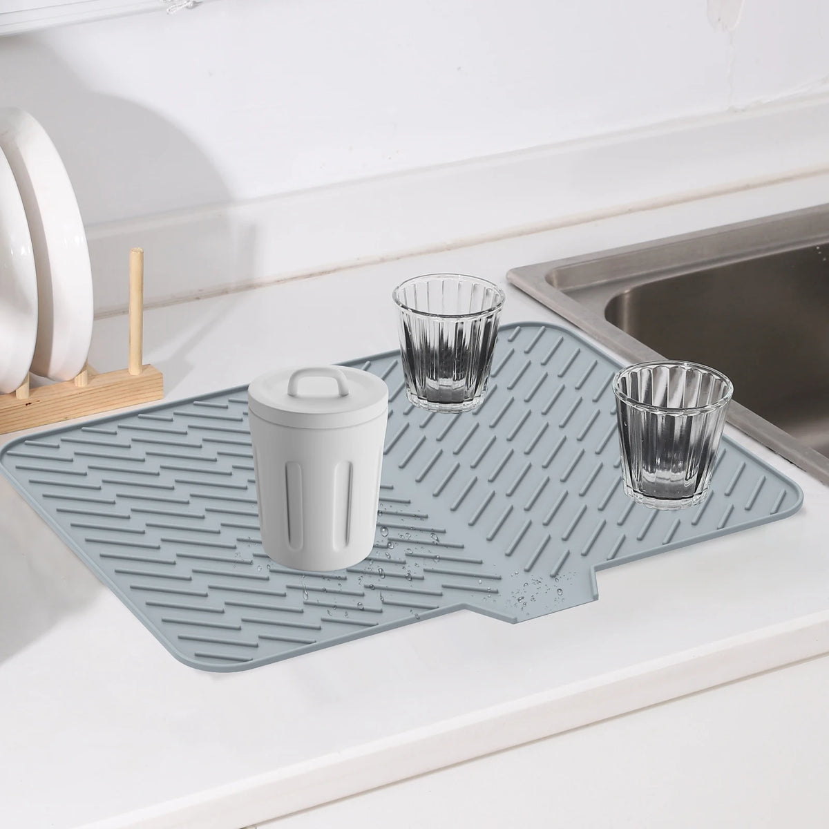 A silicone dish drying mat with a drainage ramp is placed on a sink next to drinking glasses and a storage container.