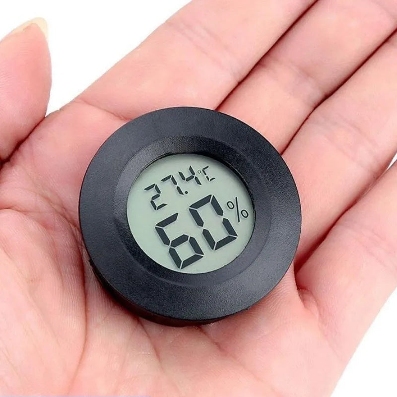 A hand holds a black mini digital hygrometer thermometer displaying 27.4°C and 60% humidity.