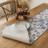 A beige linen table runner with a blue floral pattern laid on a wooden table next to a bowl of grapes.