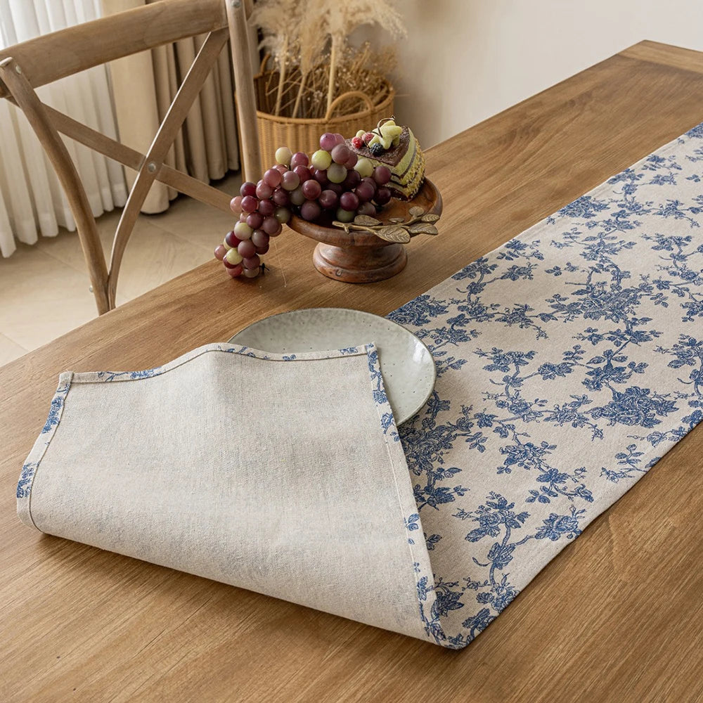 A beige linen table runner with a blue floral pattern laid on a wooden table next to a bowl of grapes.