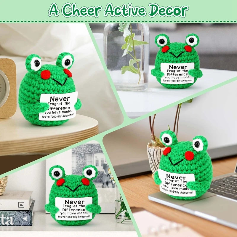Handmade crochet frog doll in green with a positive message card on a wooden surface.