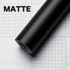 A roll of matte black carbon fiber vinyl wrap film lying on a white grid surface.