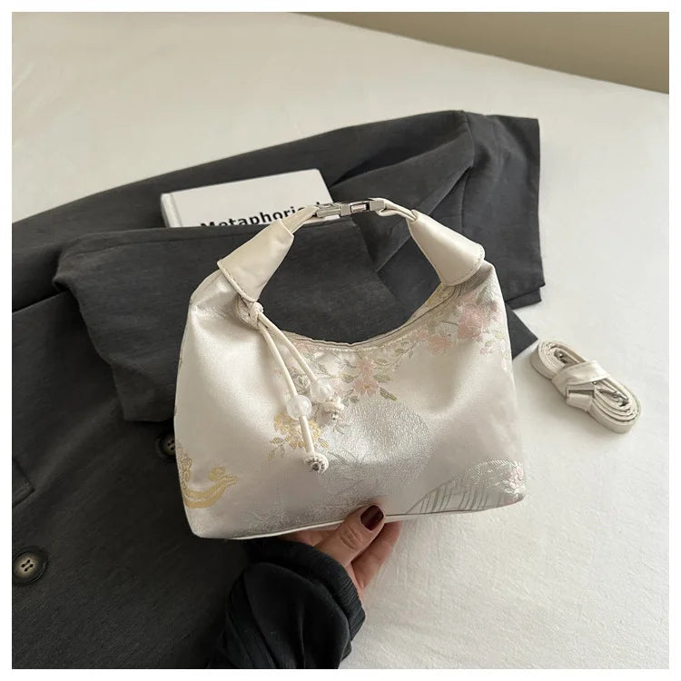 Cremiges Seiden-Crossbodytasche mit Perlenstickerei vor einem dunkelgrauen Blazer.